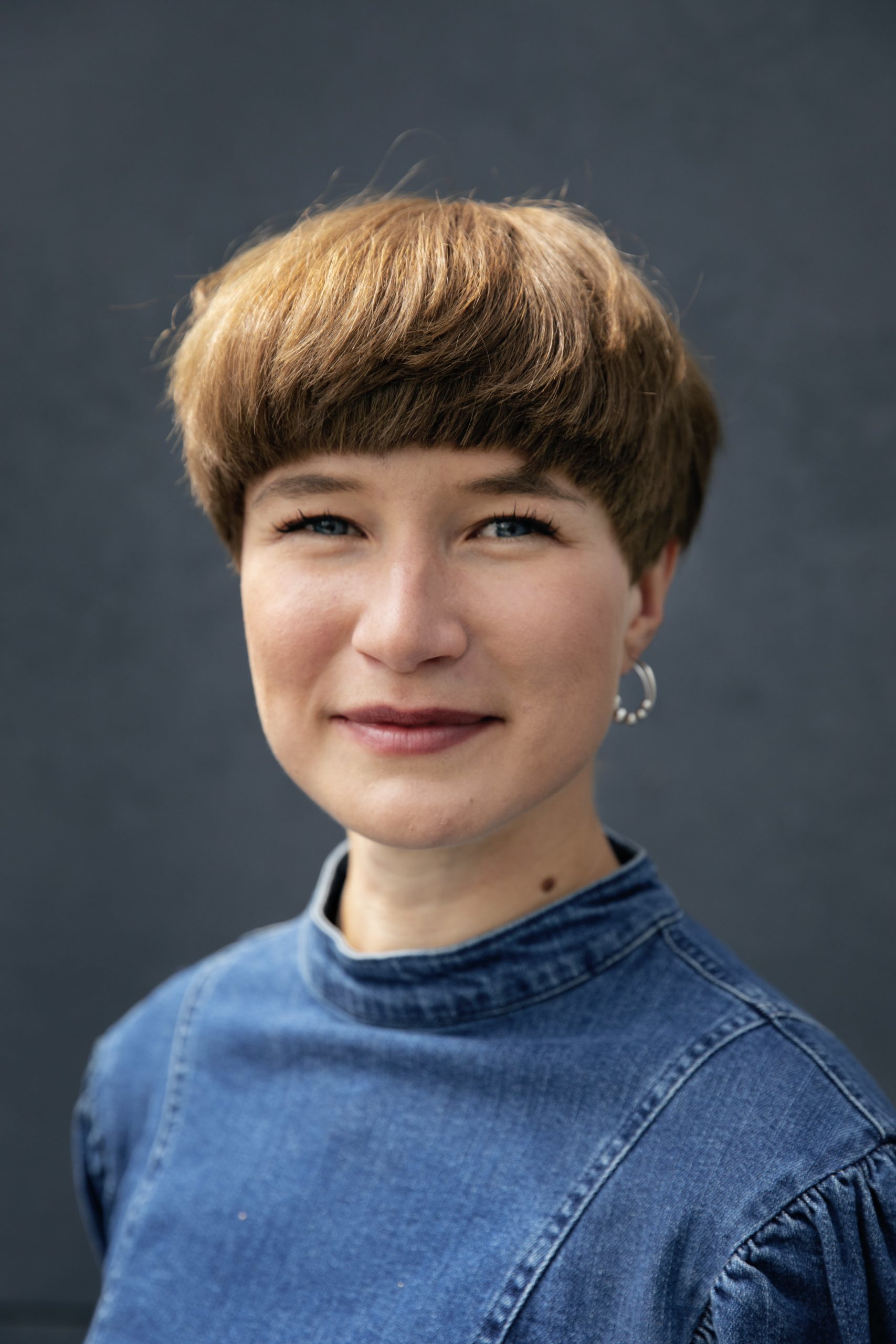 Portrait of Nikoline on a black background. She is facing the camera straight on and wearing a denim high-neck dress and silver hoop earrings.